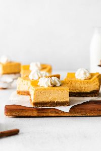 Gluten Free Pumpkin Cheesecake Bars on a parchment paper lined wood cutting board topped with whipped cream