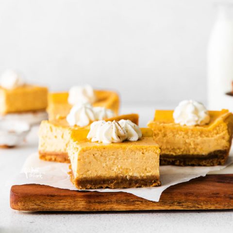 Gluten Free Pumpkin Cheesecake Bars on a parchment paper lined wood cutting board topped with whipped cream
