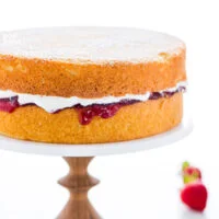 a gluten free Victoria Sponge Cake made of 2 layers of vanilla sponge cake filled with strawberry jam and whipped cream, on top of a white marble cake stand with a wood base