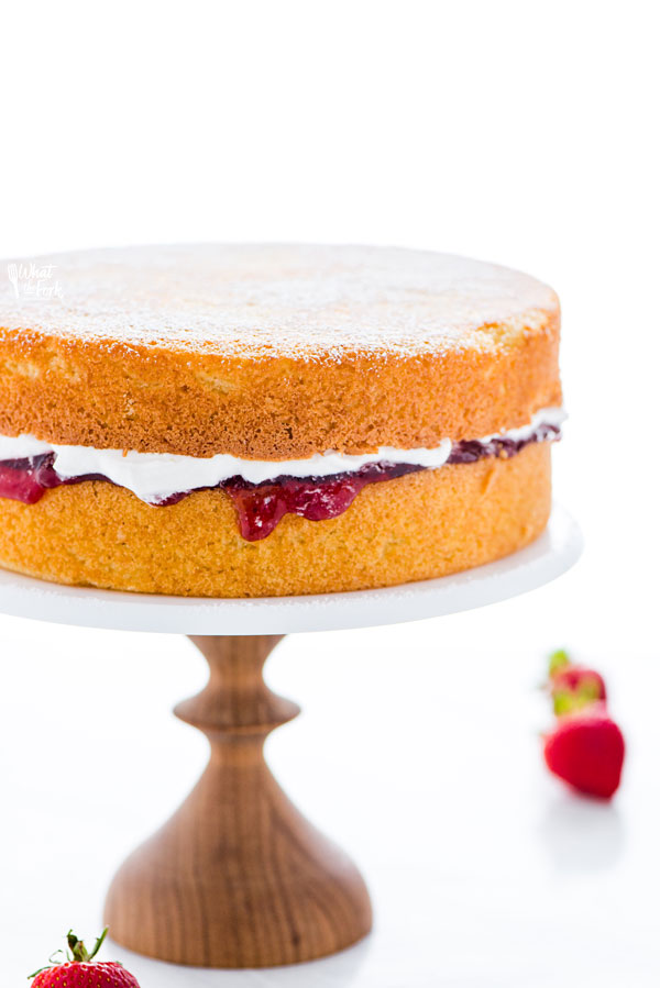 a gluten free Victoria Sponge Cake made of 2 layers of vanilla sponge cake filled with strawberry jam and whipped cream, on top of a white marble cake stand with a wood base
