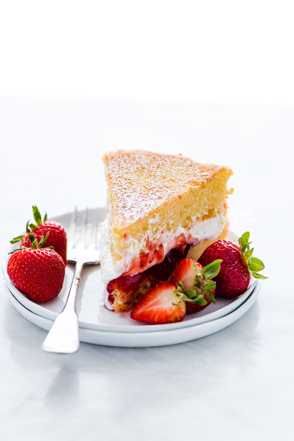 a slice of gluten free Victoria Sponge Cake on a stack of 2 small white plates with a silver fork garnished with fresh strawberries