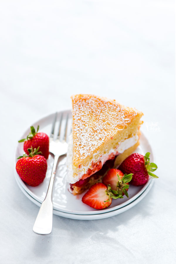 a slice of Gluten Free Victoria Sponge Cake on a white plate with a silver fork and garnished with fresh strawberries
