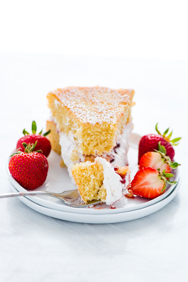 a slice of Gluten Free Victoria Sponge Cake on a stack of 2 white plates with a forkful of cake on the plate that's garnished with fresh strawberries