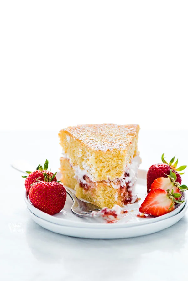 a slice of Gluten Free Victoria Sponge Cake on a stack of 2 white plates garnished with fresh strawberries that has had a forkful of cake removed from the slice