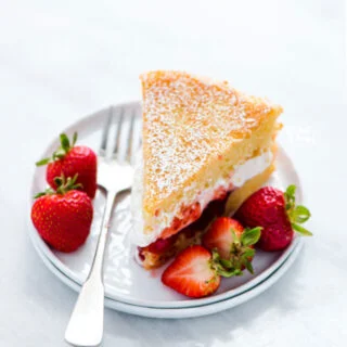 a slice of Gluten Free Victoria Sponge Cake on a white plate with a silver fork and garnished with fresh strawberries