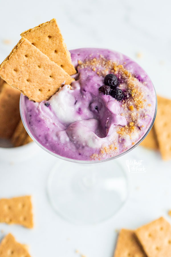 Blueberry Cheesecake Dip in a vintage martini glass garnished with crushed graham cracker crumbs, frozen blueberries, and pieces of graham cracker for serving