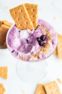 Blueberry Cheesecake Dipin a vintage martini glass garnished with crushed graham cracker crumbs, frozen blueberries and 2 pieces of graham cracker