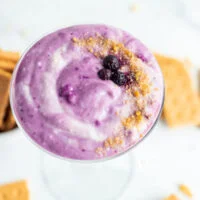 blueberry cheesecake dip in a vintage martini glass garnished with crushed graham cracker crumbs and frozen blueberries
