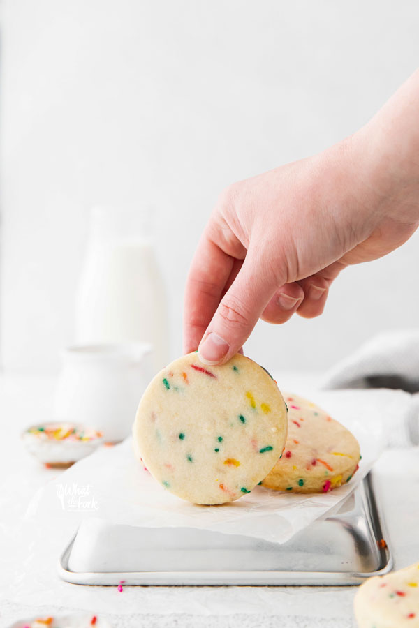 straight on shot of Gluten Free Shortbread Cookies with Sprinkles on a white parchment paper lined quarter sheet pan with a hand taking a cookie