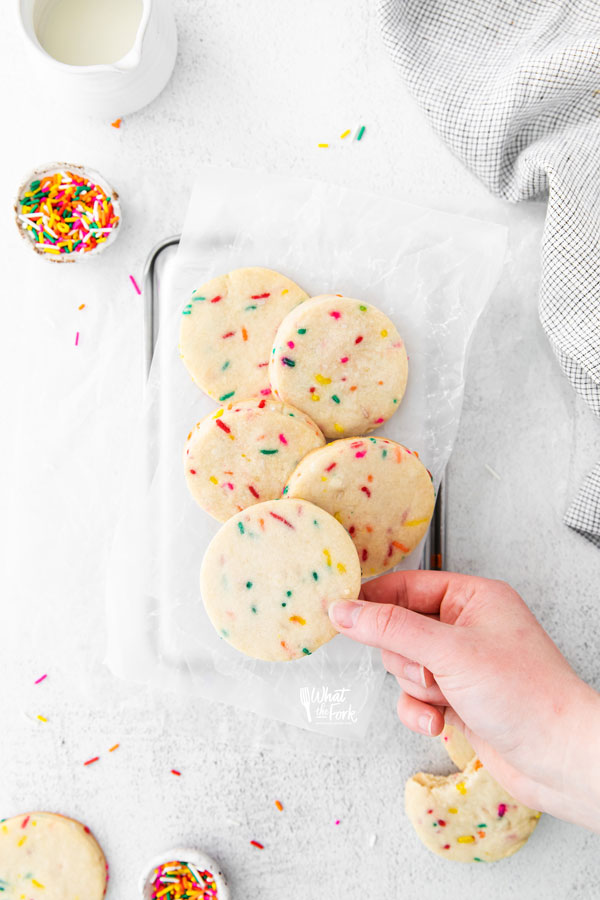 overhead shot of Gluten Free Shortbread Cookies with Sprinkles on a white parchment paper lined quarter sheet pan with a hand taking a cookie