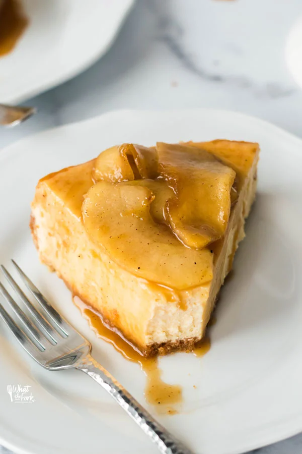 a slice of gluten free caramel apple cheesecake on a white plate with a silver fork that has a bite taken out and caramel drizzled on the plate