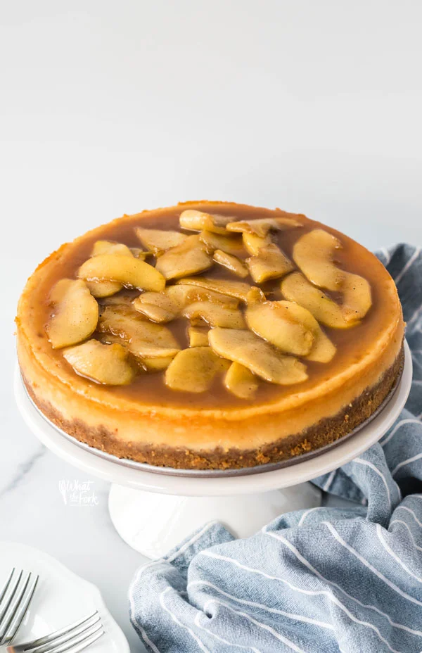 Gluten Free Caramel Apple Cheesecake on a white cake stand with a blue kitchen towel underneath