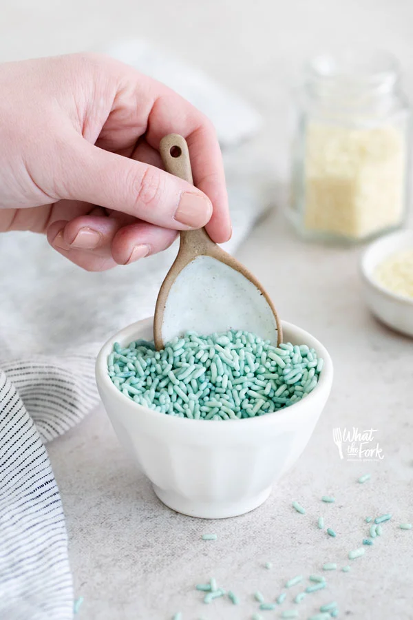 blue sprinkles in a small white bowl with a hand holding a brown and white spoon starting to scoop the sprinkles