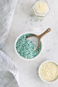 overhead shot of blue sprinkles in a white bowl with a brown and white spoon with a bowl of white sprinkles on the side