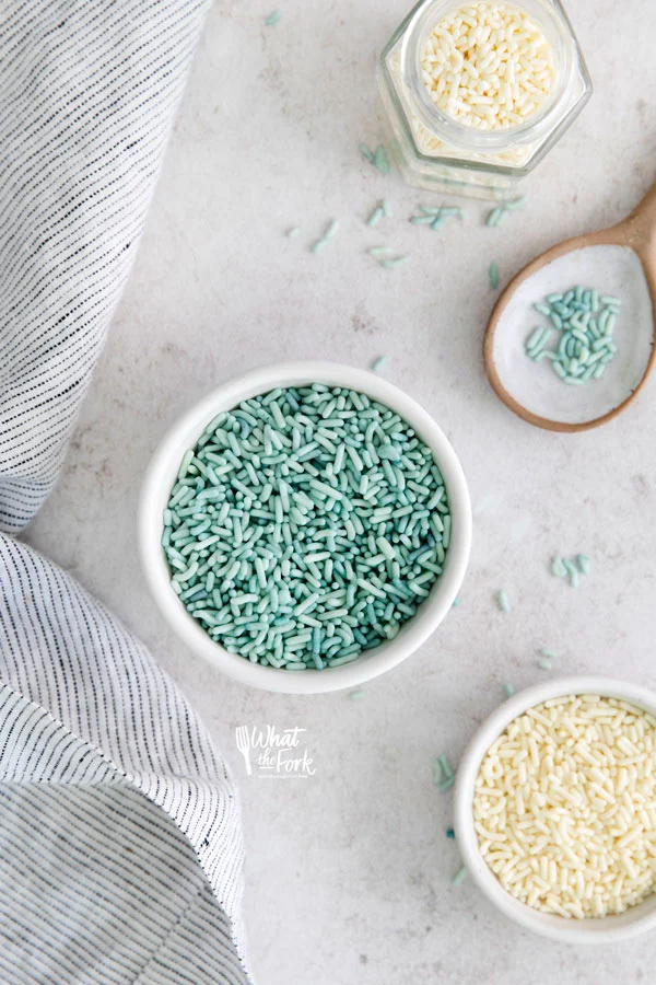 overhead shot of blue sprinkles in a white bowl with a brown and white spoon on the side and a small bowl of white sprinkles in the bottom right hand corner