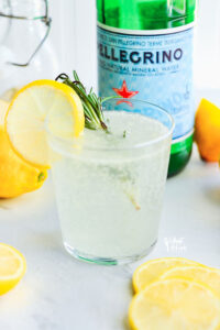 Non-Alcoholic Rosemary Citrus Spritzers in a glass garnished with a lemon wheel on the side of the glass and a sprig of rosemary in the glass. A bottle of San Pellegrino is in the background