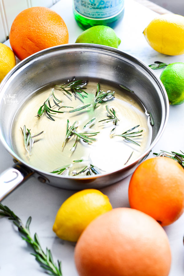 simple syrup in a metal pan with sprigs of rosemary in it. oranges, lemons, and limes surround the pan