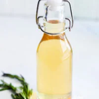 learn how to make simple syrup with rosemary! This image shows a bottle of rosemary simple syrup with a sprig of rosemary next to it.