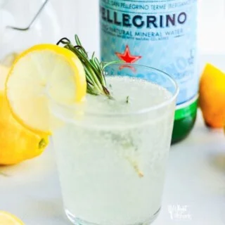 Non-Alcoholic Rosemary Citrus Spritzers in a glass garnished with a lemon wheel on the side of the glass and a sprig of rosemary in the glass. A bottle of San Pellegrino is in the background