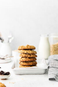 a stack of gluten free oatmeal raisin cookies piled high on a small silver metal sheet pan lined with white parchment paper with a glass of milk in the background