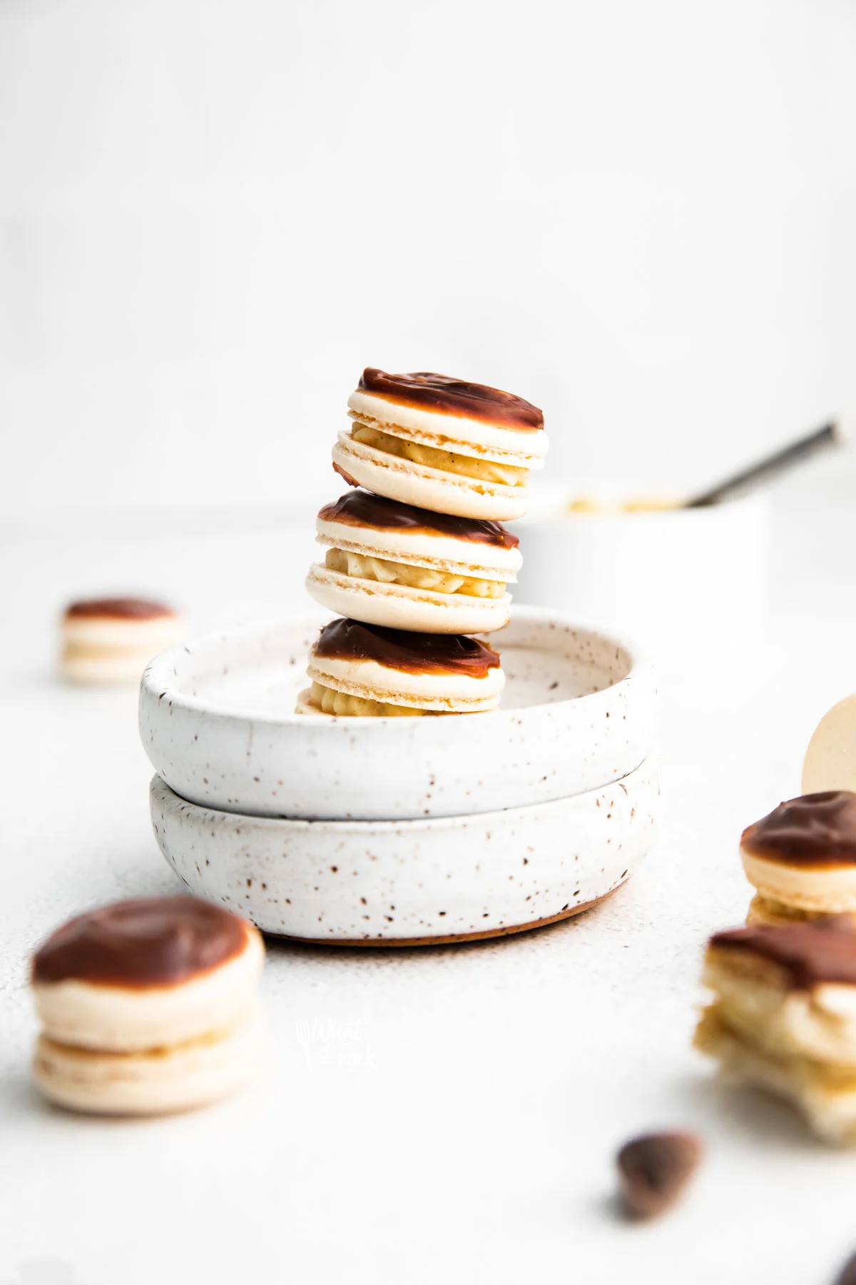 a stack of Boston Cream Pie Macarons in a small white bowl with more macarons on the surface diagonally in front