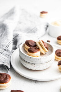 three Boston Cream Pie Macarons in a small pottery bowl on top of a plate with more macarons and a bowl of pastry cream and macarons scattered in the background