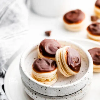 three Boston Cream Pie Macarons in a small pottery bowl on top of a plate with more macarons and a bowl of pastry cream in the background