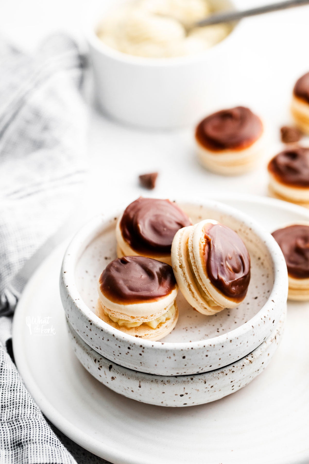 three Boston Cream Pie Macarons in a small pottery bowl on top of a plate with more macarons and a bowl of pastry cream in the background
