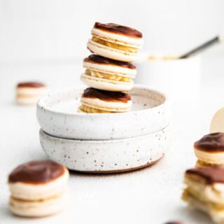 a stack of Boston Cream Pie Macarons in a small white bowl with more macarons on the surface diagonally in front