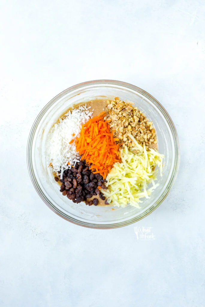 overhead shot of mixed gluten free morning glory muffins batter in a clear glass bowl with nuts, shredded carrots, grated apples, raisins, and shredded coconut on top ready to be mixed in.