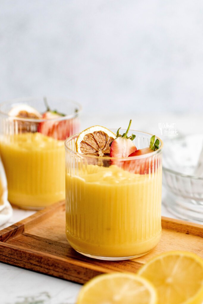 cooked lemon pudding recipe in 2 rimmed glasses topped with charred lemon wheels and a strawberry sliced in half