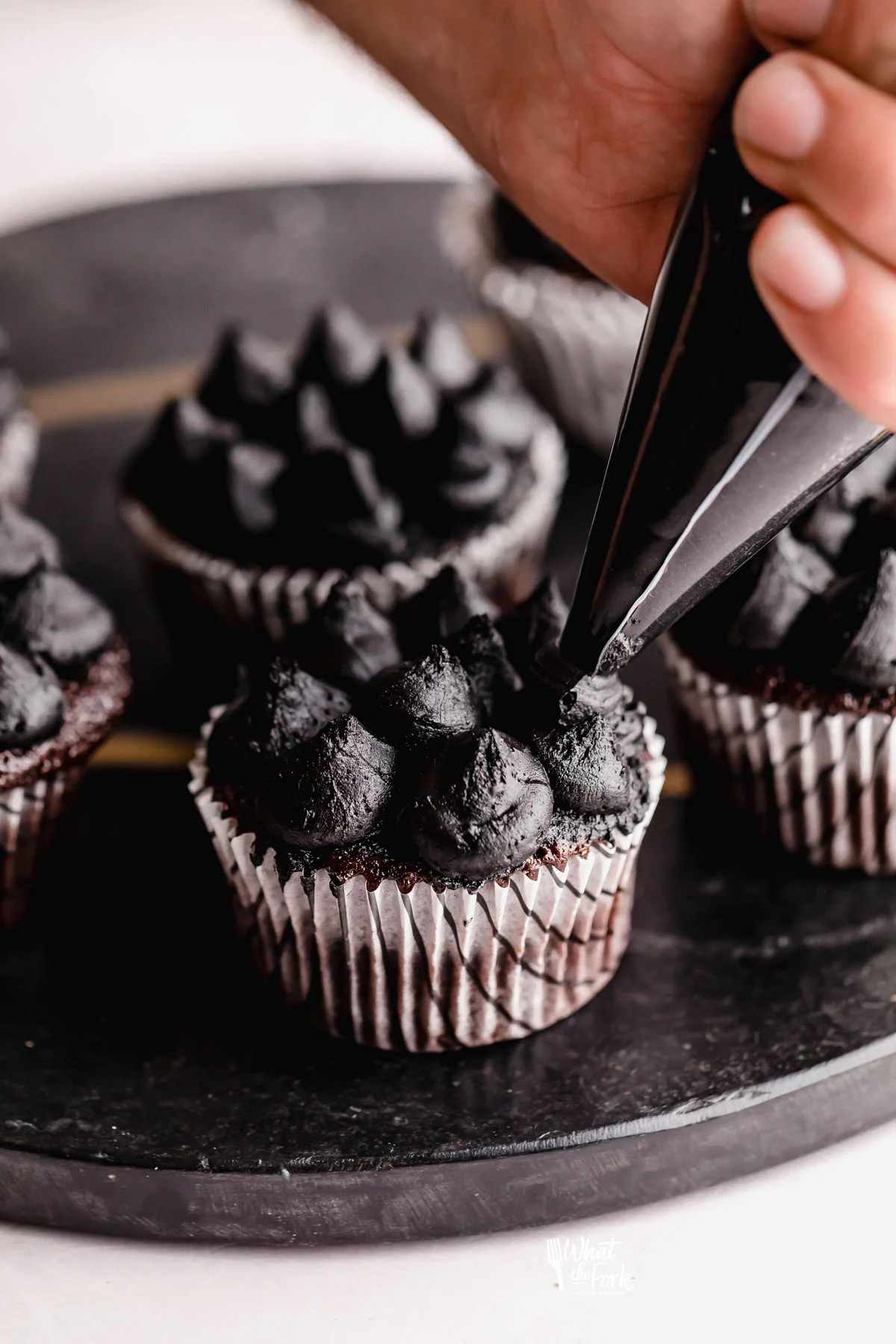 a hand holding a clear piping bag filled with black frosting piping dollops of it onto a chocolate cupcake in a white paper liner