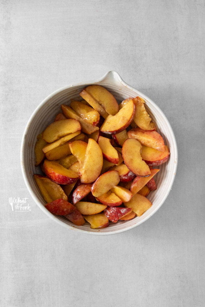 a large white bowl of fresh sliced peaches tossed with sugar, cornstarch, and cinnamon to make gluten free peach cobbler