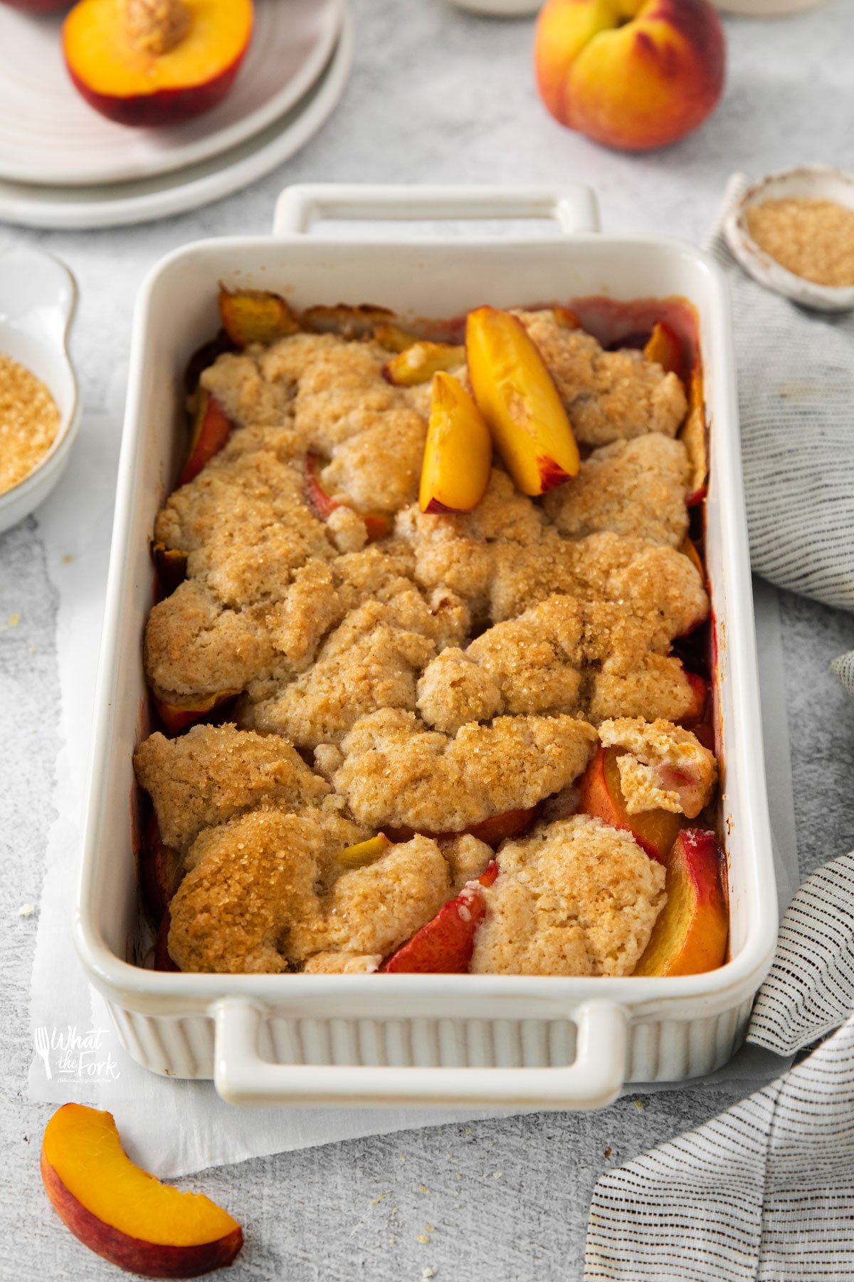 baked gluten free peach cobbler recipe in a white rectangular baking dish with handles on a white surface surrounded by fresh peaches and peach slices