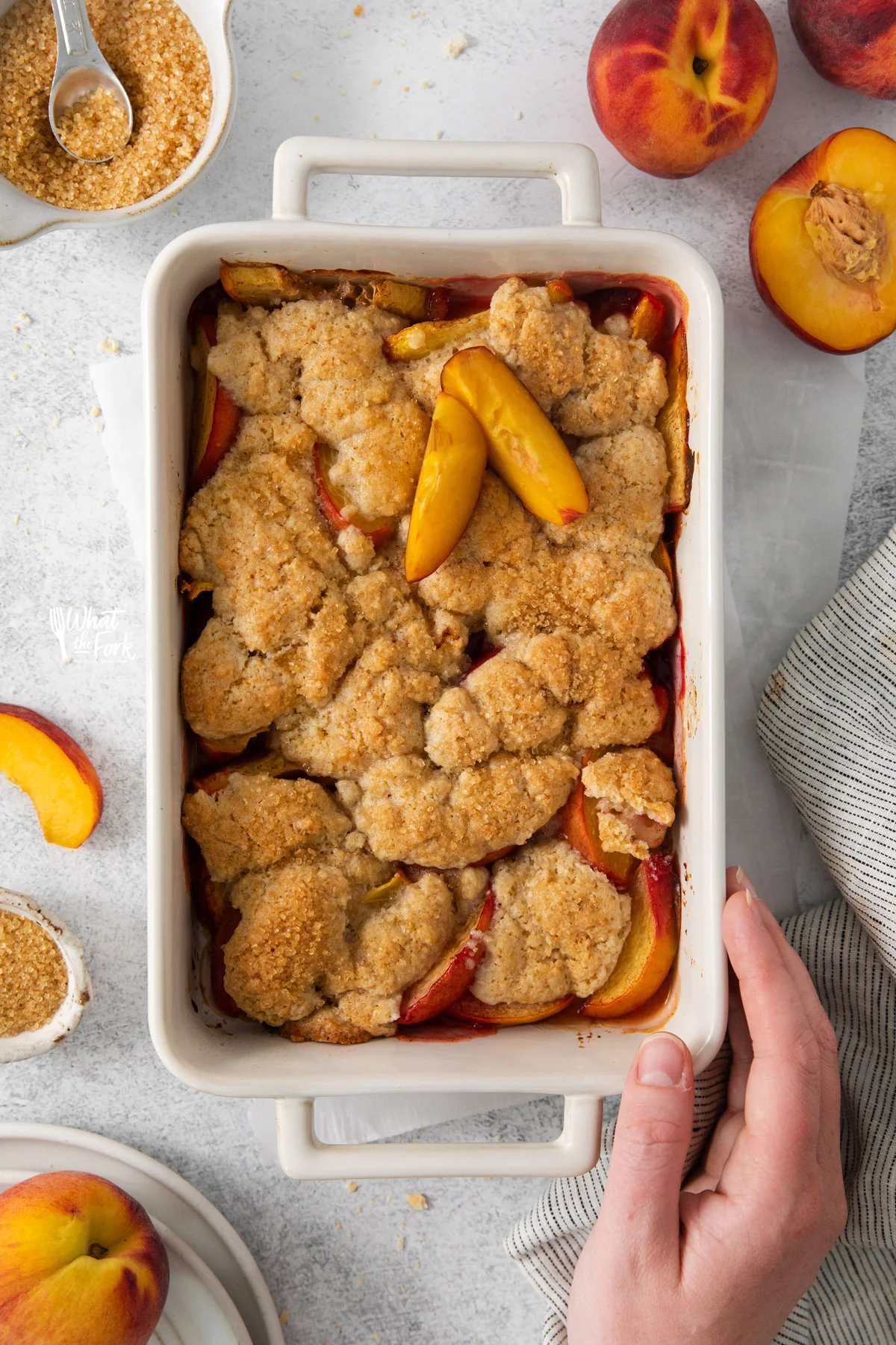 a hand on a dish of gluten free peach cobbler in a white rectangle baking dish