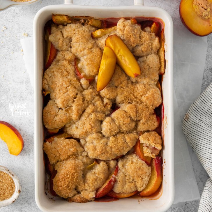 overhead shot of a baked gluten free peach cobbler recipe in a white rectangular baking dish with handles on a white surface surrounded by fresh peaches and peach slices