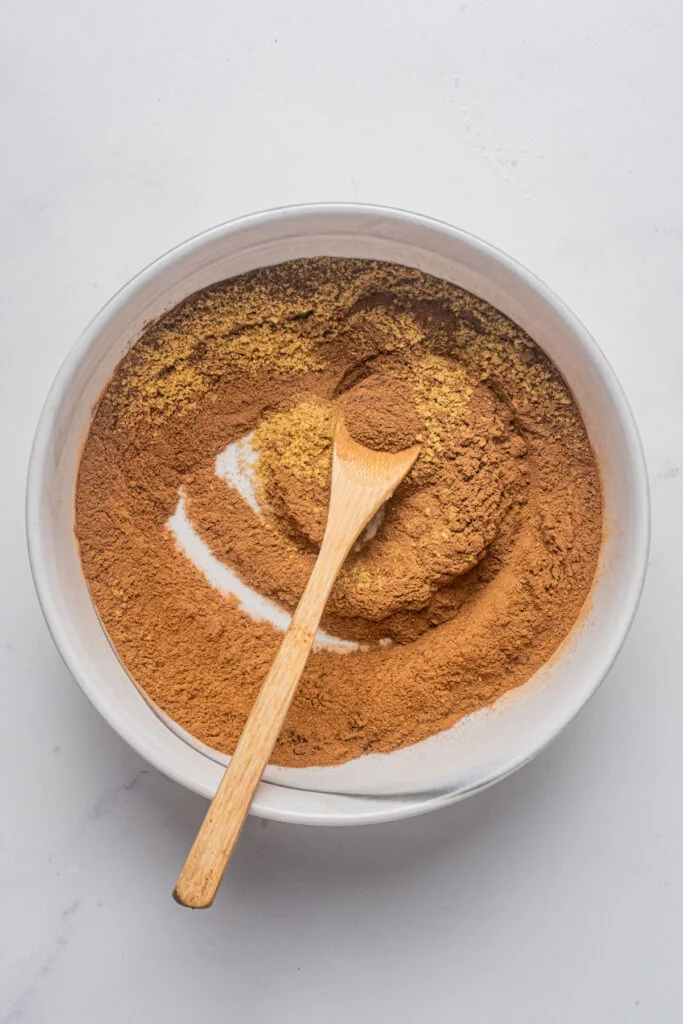 ingredients for homemade pumpkin pie spice in a small white bowl that have been mixed together with a small wooden spoon
