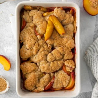 overhead shot of a baked gluten free peach cobbler recipe in a white rectangular baking dish with handles on a white surface surrounded by fresh peaches and peach slices