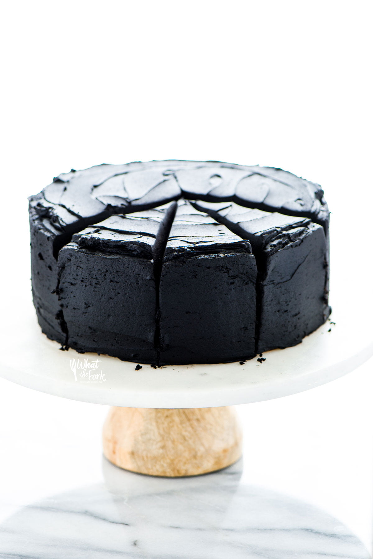 a baked and frosted gluten free black velvet cake recipe on top of a white marble cake stand with a wood base. It's been frosted with black buttercream and has 3 slices cut but not served.