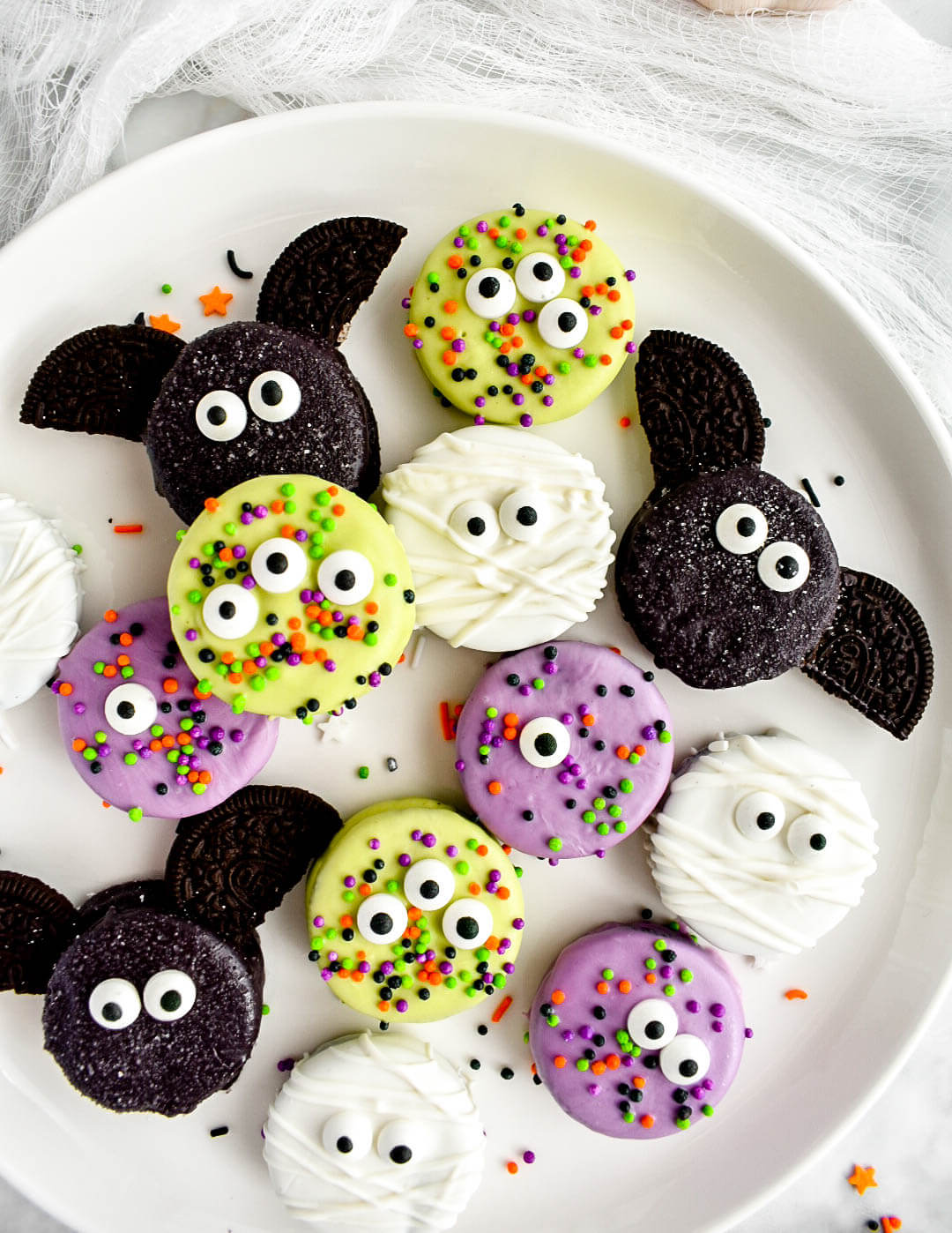 Halloween Chocolate Covered Oreos are decorated to look like green monsters, mummies, bats, and purple monsters and are arranged on a white plate.