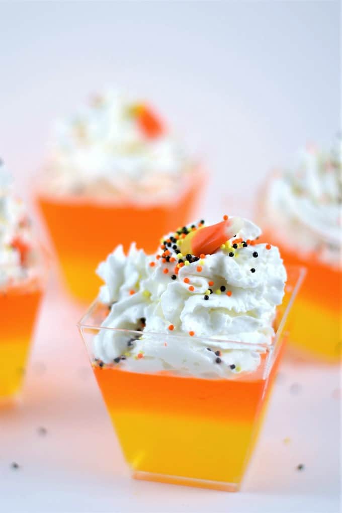 candy corn jello cups are made with layers of lemon and orange jello. They're topped with whipped cream, Halloween sprinkles, and a candy corn.
