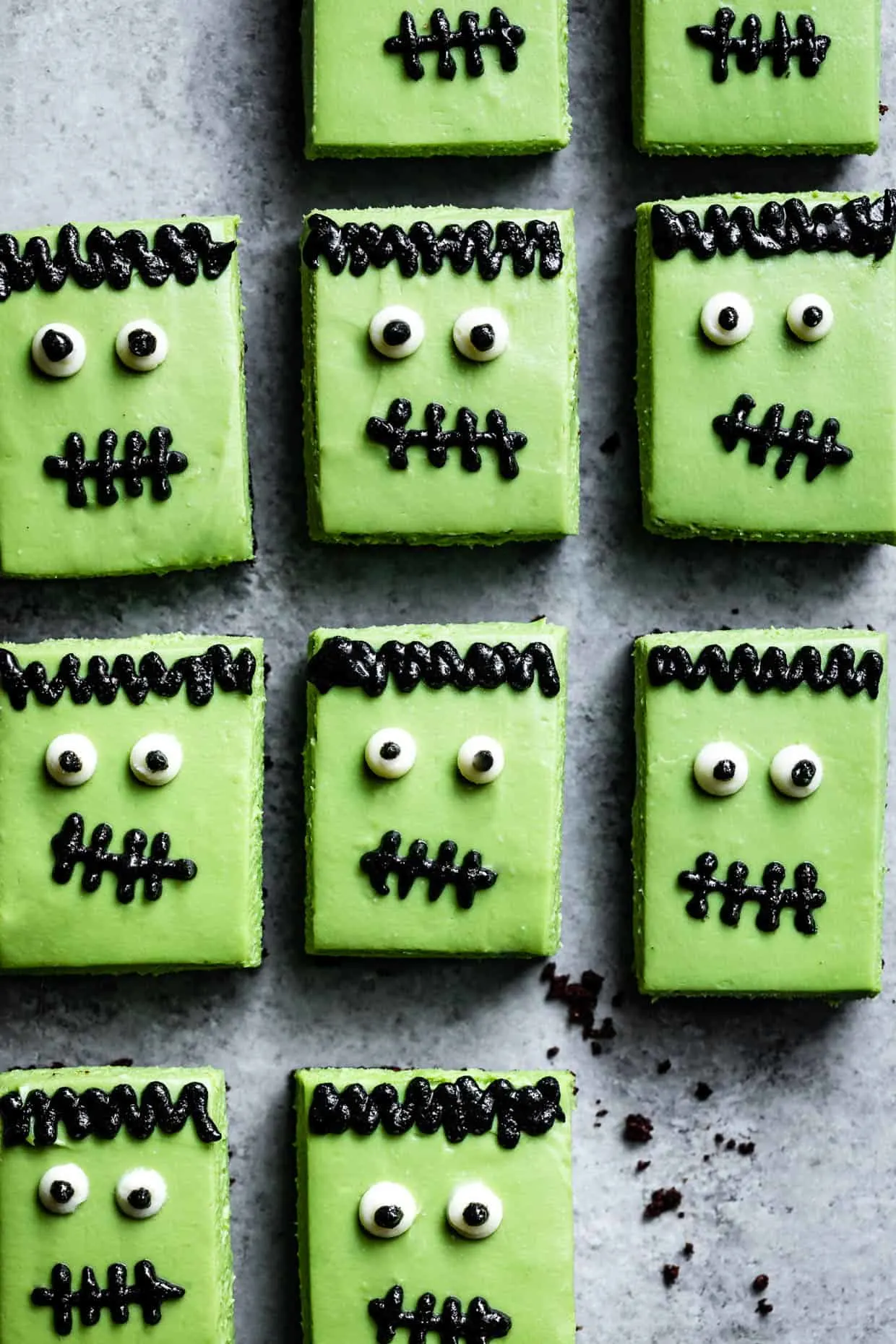 Frankenstein Cheesecake Bars lined up on a gray surface - they're decorated with black sesame paste and candy eyes