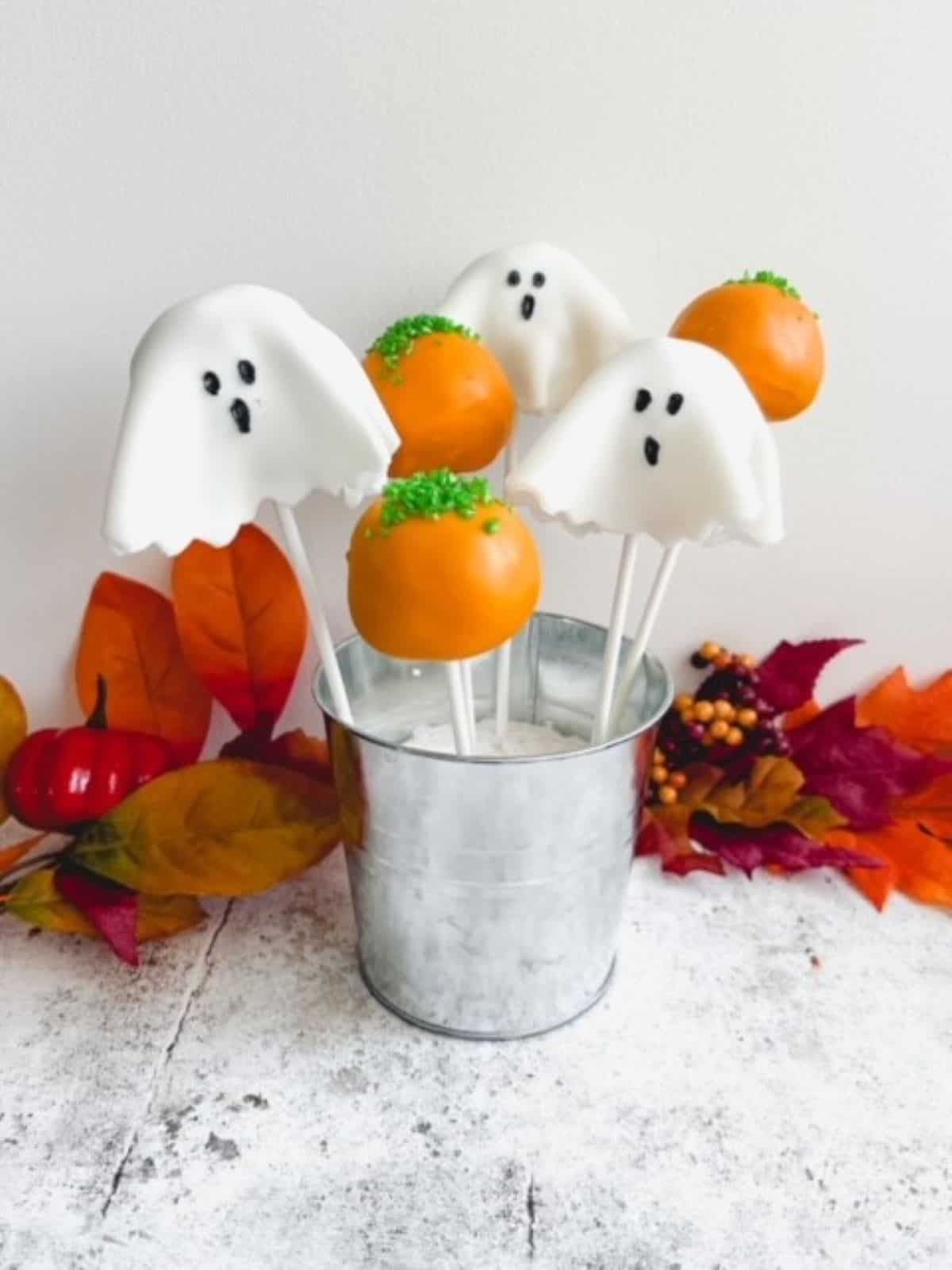 Halloween Cake Pops decorated like pumpkins and ghosts in a silver pail with artificial leaves in the background