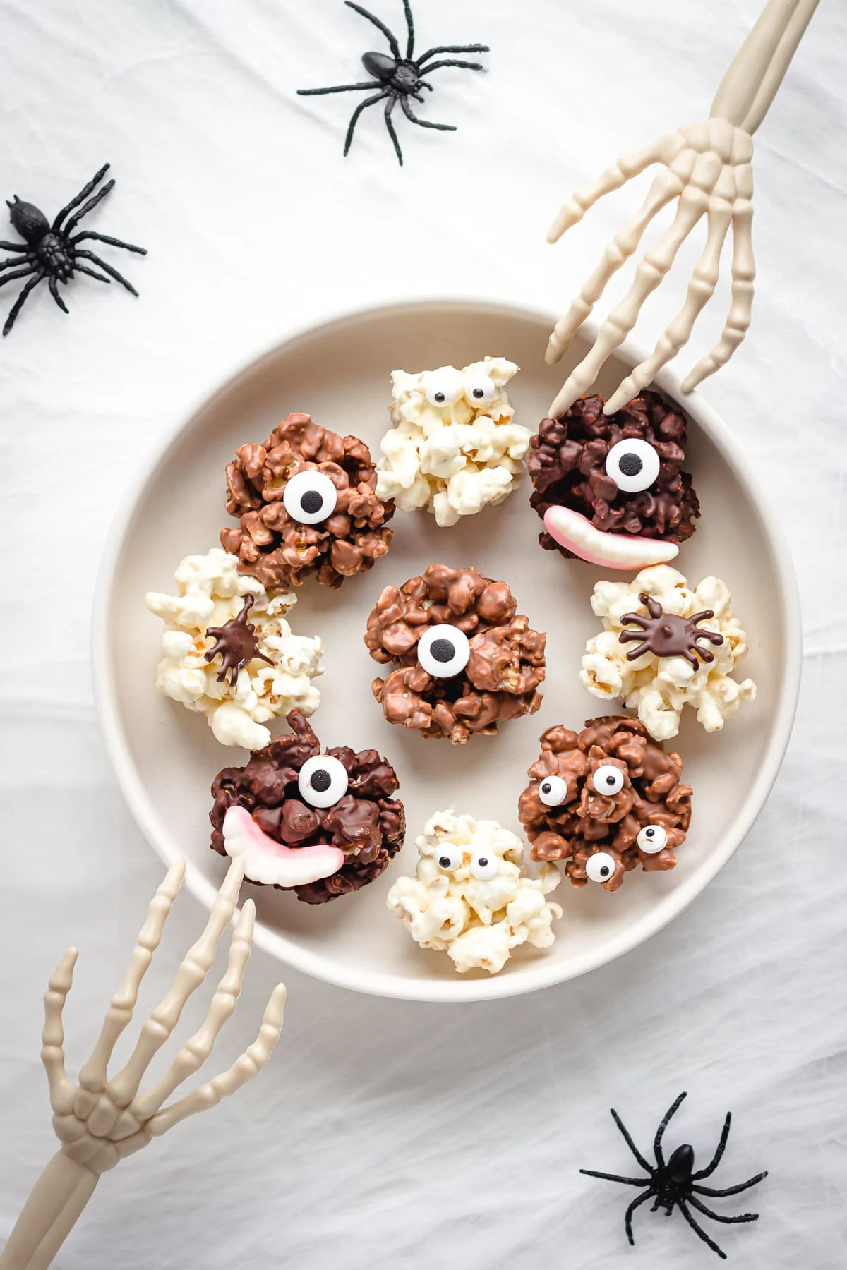 Halloween Popcorn Balls decorated with candy eyes and creepy teeth on a white plate with two skeleton hands reaching for them.