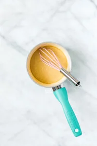 maple glaze in a small white saucepan with a aqua blue handle with a silver and pink whisk in it