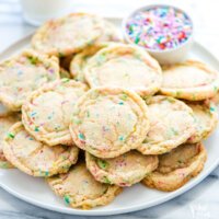 a large, round, white plate full of chewy gluten free sugar cookies with sprinkles with a small bowl of rainbow sprinkles on the side with a small glass of milk in the background