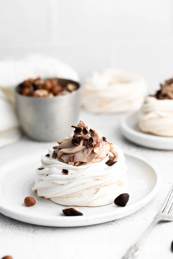 an individual Nutella Pavlova on a round white plate filled with Nutella Whipped Cream and topped with toasted hazelnuts and chocolate shavings.