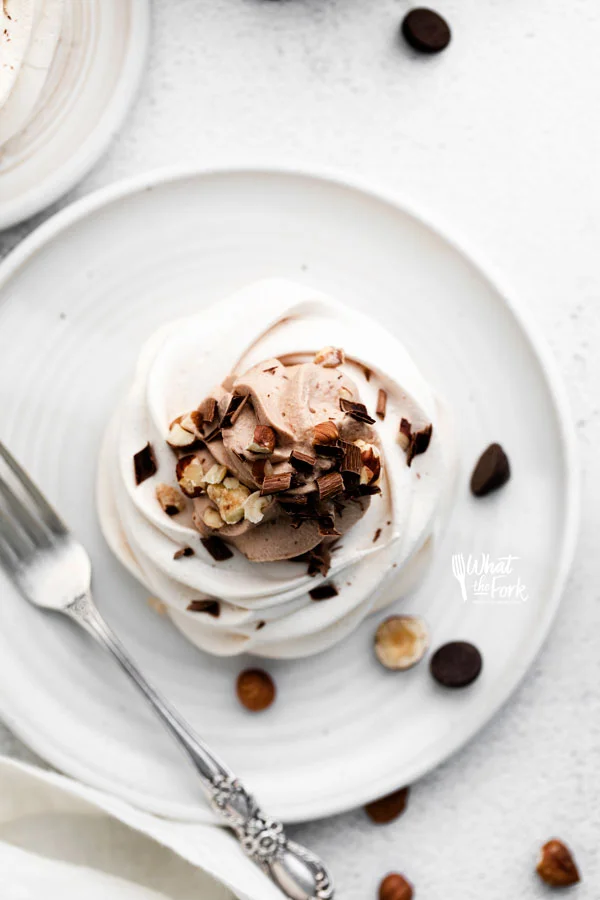 overhead shot of an Individual Nutella Pavlova on a round white plate with a silver spoon garnished with chopped hazelnuts and chocolate