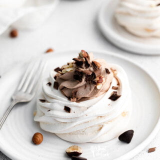 Nutella pavlovas plated on a round white plate with a silver spoon. The individual pavlova is filled with Nutella whipped cream and topped with chopped hazelnuts and chocolate.