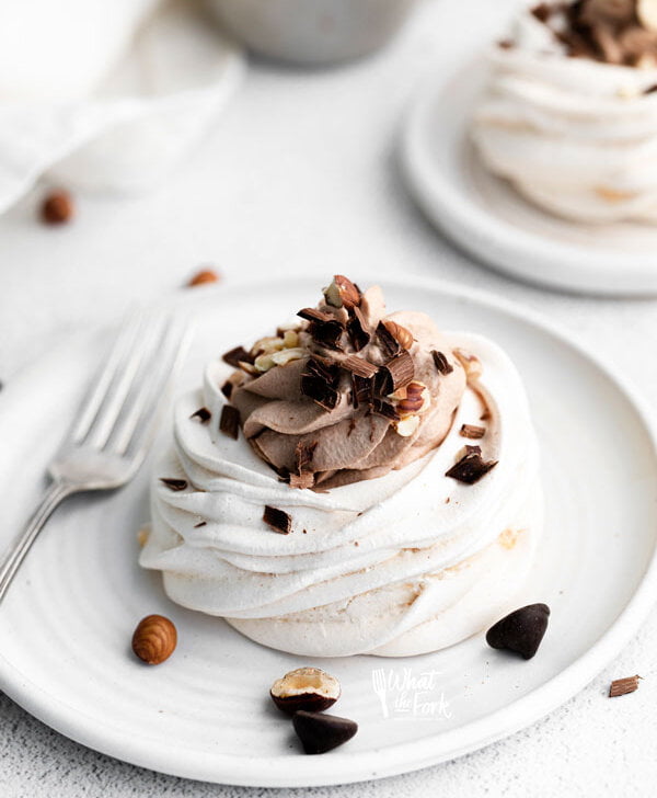 Nutella pavlovas plated on a round white plate with a silver spoon. The individual pavlova is filled with Nutella whipped cream and topped with chopped hazelnuts and chocolate.